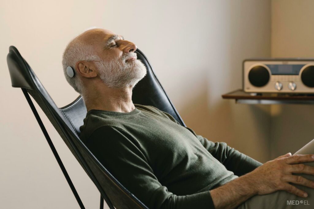 Ein älterer Mann mit einem Cochlea Implantat sitzt in einem Liegestuhl und im Hintergrund steht ein Radio.